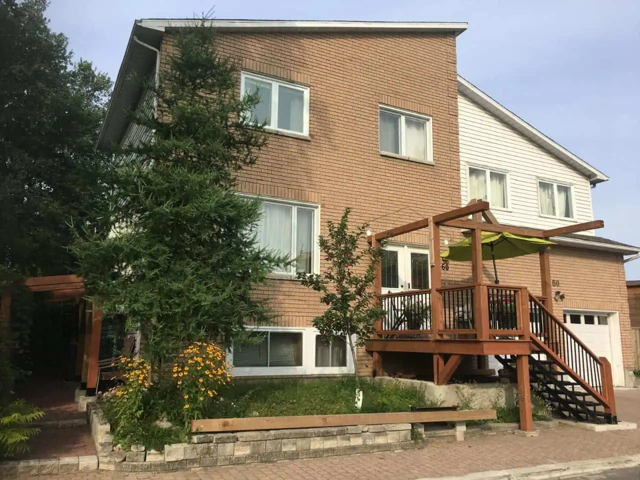 This charming two-story house, adorned with brick and white siding, features a wooden deck with a pergola and green shade cover. The small front garden is filled with vibrant flowers and greenery. On the right, a garage offers convenient access to what would be an ideal location for a massage clinic.