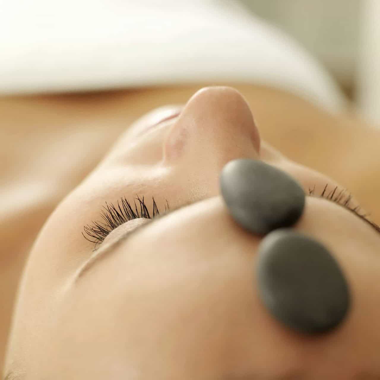 Close-up of a person with their eyes closed, lying down, indulging in a soothing hot stone massage. Two smooth, dark stones are placed on their forehead, embodying tranquility in an Ottawa spa treatment.